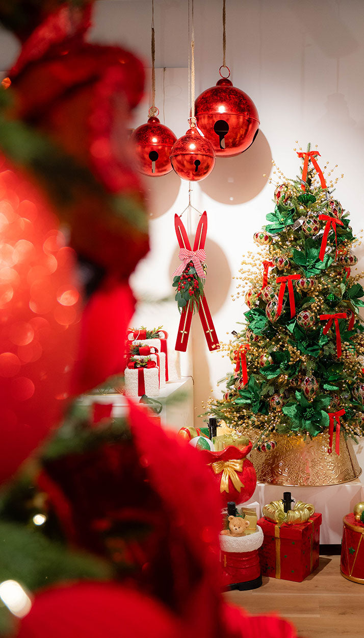 A festive Christmas tree decorated with red ribbons, gold ornaments, and a gold tree skirt, with red hanging ornaments and wrapped gifts on a light-colored floor. The foreground is blurred with large red bokeh baubles.